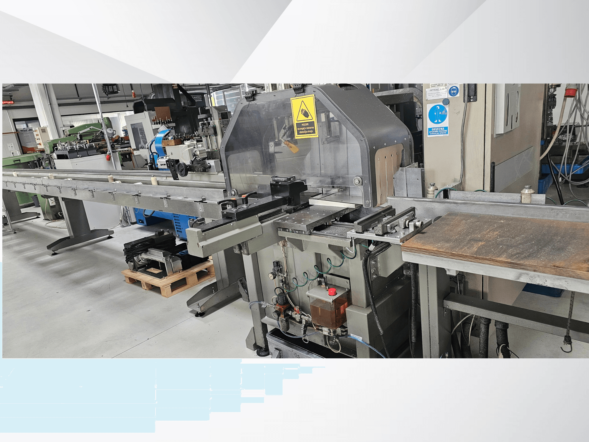 Industriële snijmachine in zijaanzicht, met een lange transportband, bedieningspaneel en veiligheidswaarschuwingen.