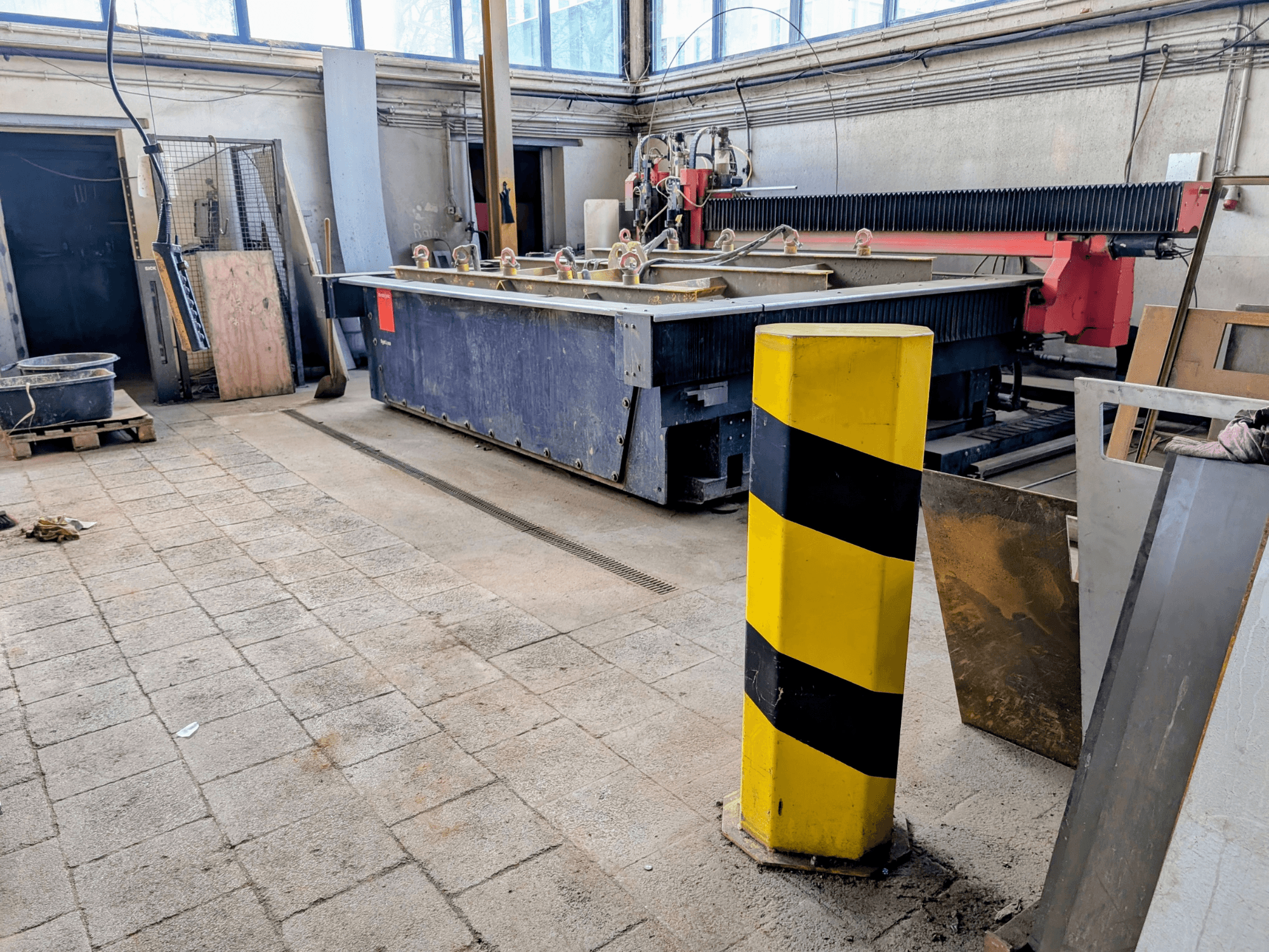 Bystronic Byjet L 2030 waterstraalsnijmachine in een werkplaats, zijaanzicht, met een gele veiligheidspaal en omringende materialen.
