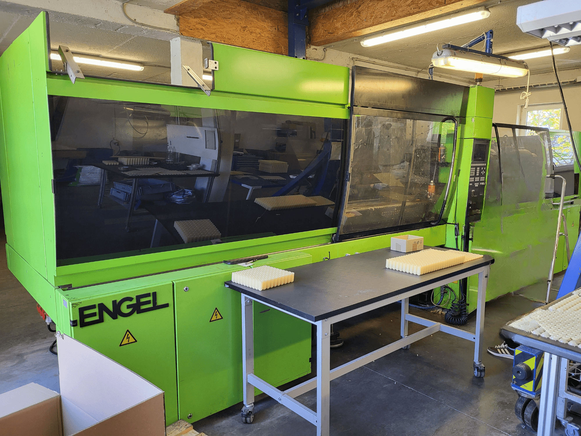 De hydraulische spuitgietmachine Green Engel ES1800/300HL, gezien vanaf de voorkant, met gestapelde materialen op een werktafel.
