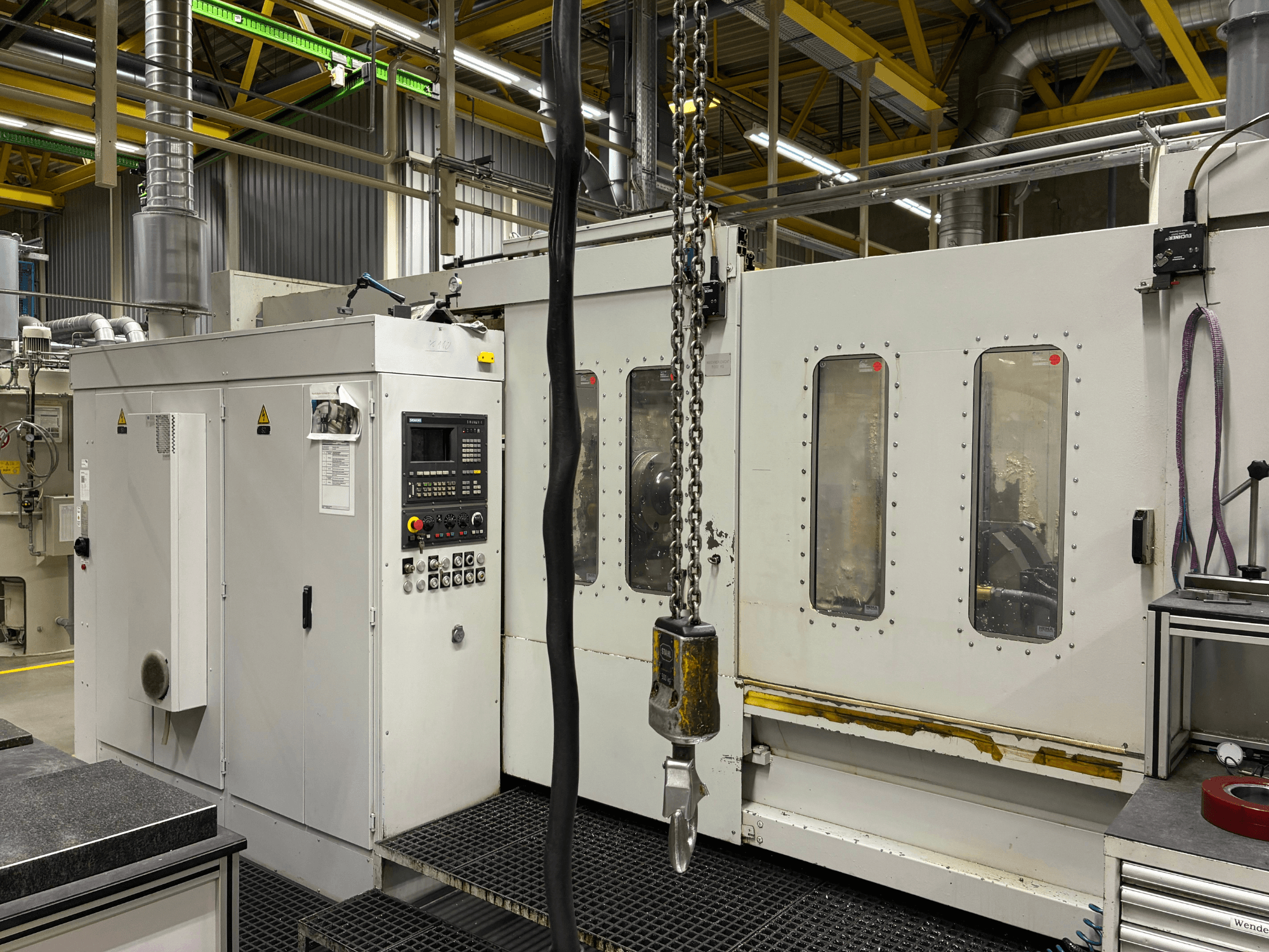 Industriële CNC machine met bedieningspaneel, zijaanzicht; met ramen, kettingen en metalen onderdelen in een fabrieksomgeving.