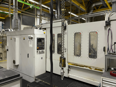 Industriële CNC machine met bedieningspaneel, zijaanzicht; met ramen, kettingen en metalen onderdelen in een fabrieksomgeving.