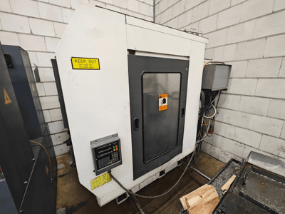 Industriële CNC machine, zijaanzicht; met bedieningspaneel, waarschuwingsborden en houten stukken op een werkblad.